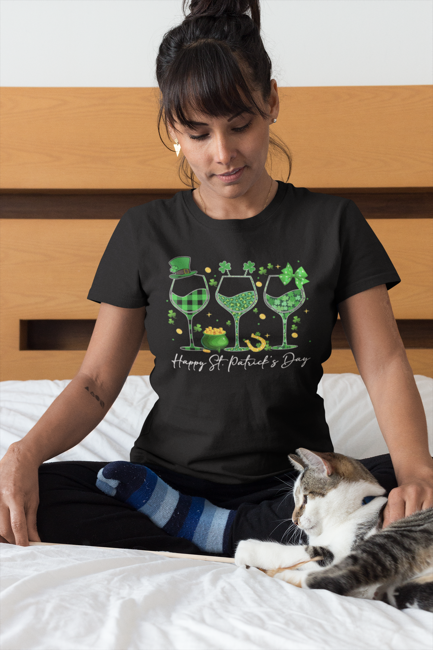 Woman wearing a black St. Patrick's Day t-shirt with green wine glasses and text, sitting on a bed with a cat.